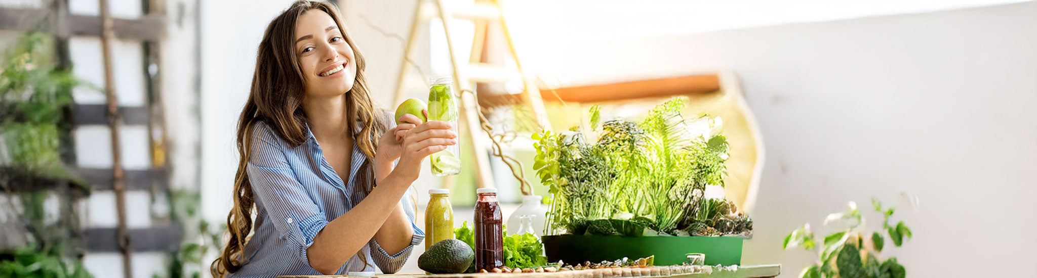 Eine lächelnde Frau sitzt an einem Tisch mit frischem Gemüse, grünen Pflanzen und Saftgläsern, in der Hand einen grünen Smoothie und einen Apfel in einem hellen, sonnendurchfluteten Raum.