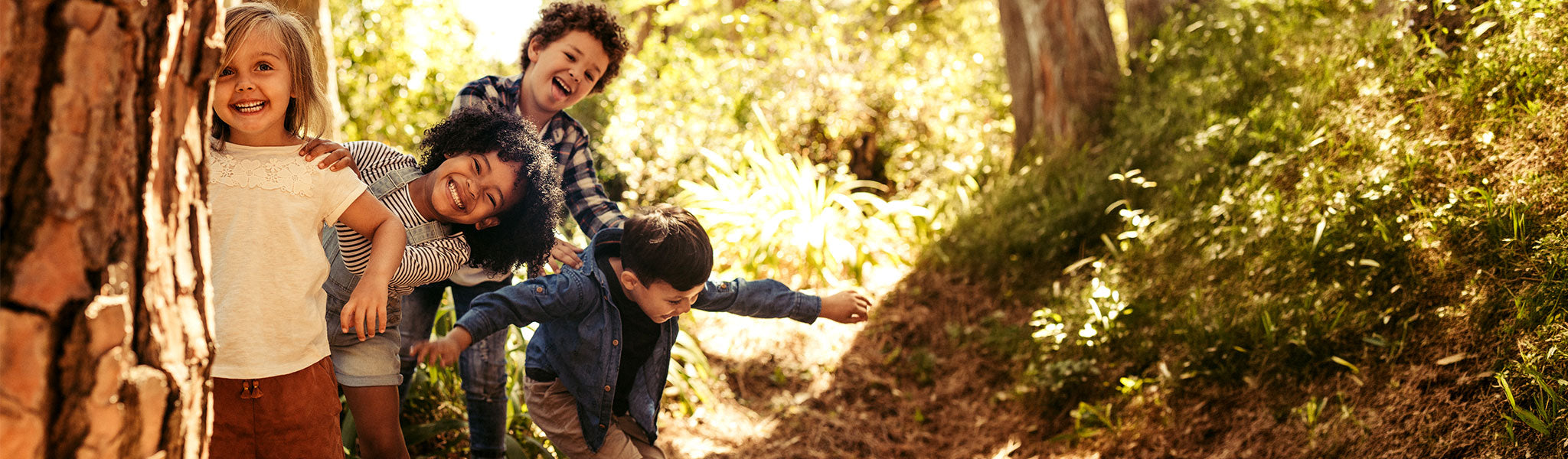 Vier kleine Kinder, die lächelnd gemeinsam im Freien spielen, in einer Reihe stehen und hinter einem Baum in einer sonnigen, bewaldeten Gegend hervorschauen.
