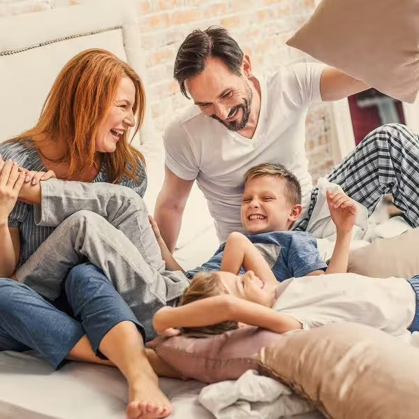 Eine fröhliche vierköpfige Familie im Pyjama lacht und spielt auf einem sonnenbeschienenen Bett – und strahlt das Wohlbefinden aus, das CitroPlus® 1200 forte (Bio) von gse.organic.supplements bietet, während sie diesen glücklichen, entspannten Moment zusammen genießt.